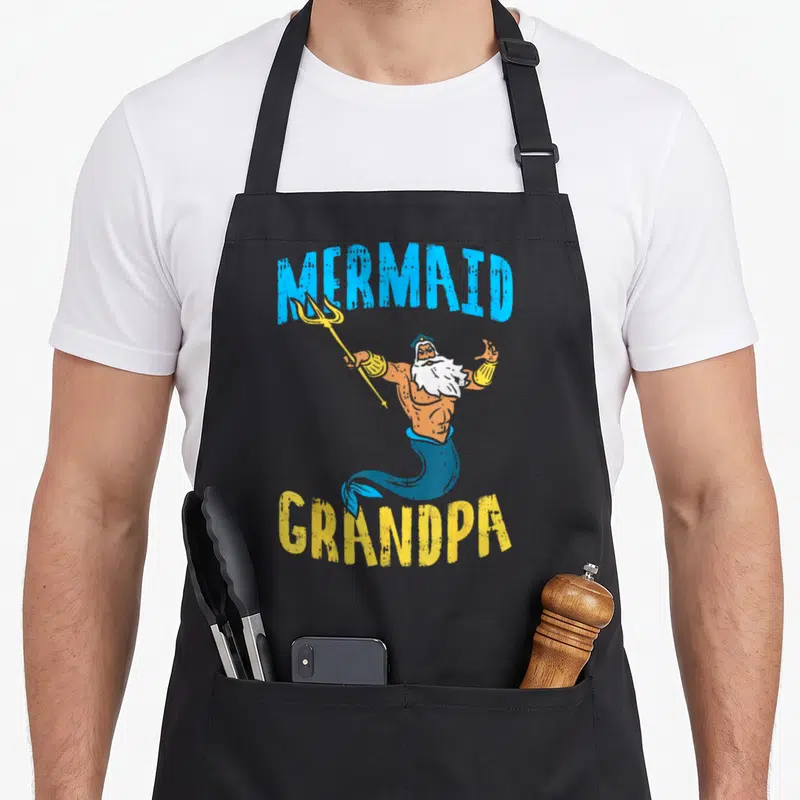 Neptune grandpa apron - Cartoon merman holding trident with blue MERMAID text above and yellow GRANDPA text below — shown on a black cotton canvas apron against a white background