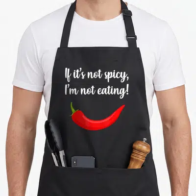 Adventure thai food apron - White script reading If it's not spicy I'm not eating above a red cartoon chili pepper — shown on a black cotton canvas apron against a white background