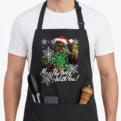 Funny christmas lights apron - Chewbacca in Santa hat tangled in red green white lights with May The Force Be With You — shown on a black cotton canvas apron against a white background