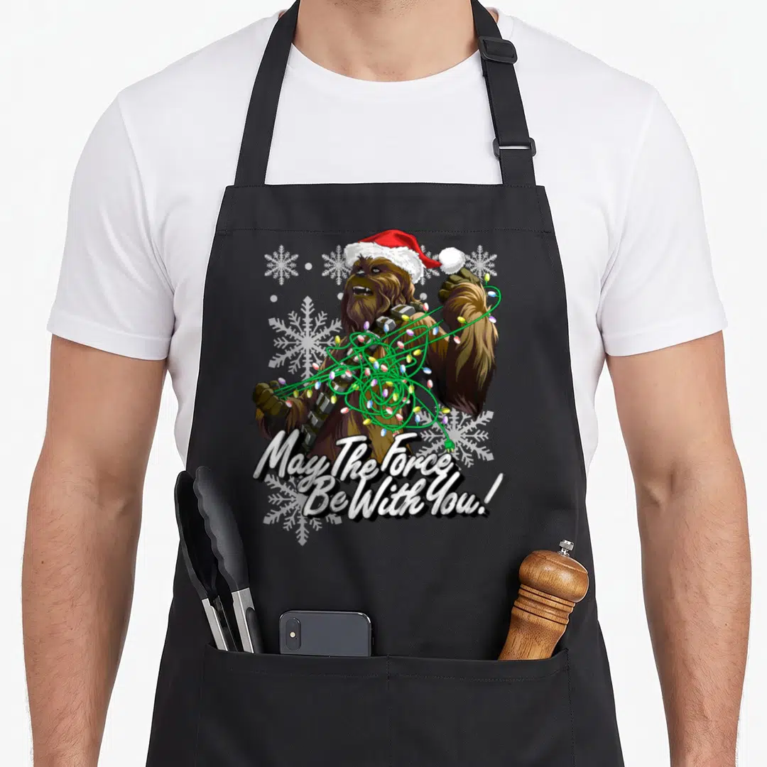 Funny christmas lights apron - Chewbacca in Santa hat tangled in red green white lights with May The Force Be With You — shown on a black cotton canvas apron against a white background