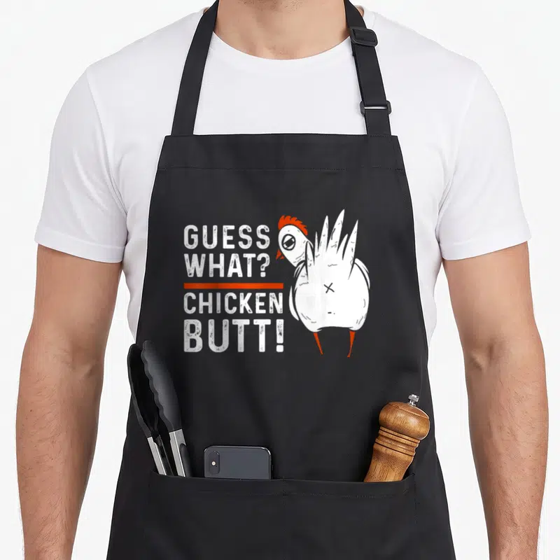 Guess chicken apron - Stacked text Guess What Chicken Butt next to cartoon chicken facing left — shown on a black cotton canvas apron against a white background
