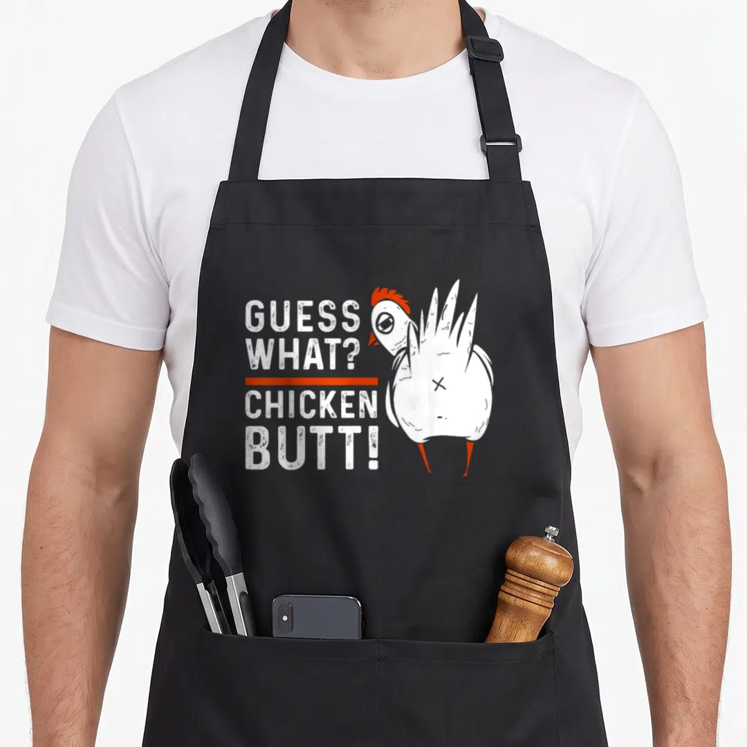 Guess chicken apron - Stacked text Guess What Chicken Butt next to cartoon chicken facing left — shown on a black cotton canvas apron against a white background