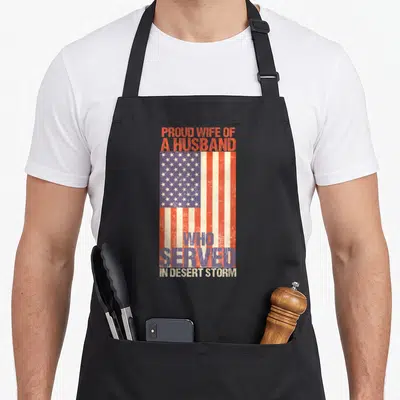 Support best husband apron - Distressed USA flag with PROUD WIFE OF A HUSBAND WHO SERVED IN DESERT STORM text in red — shown on a black cotton canvas apron against a white background