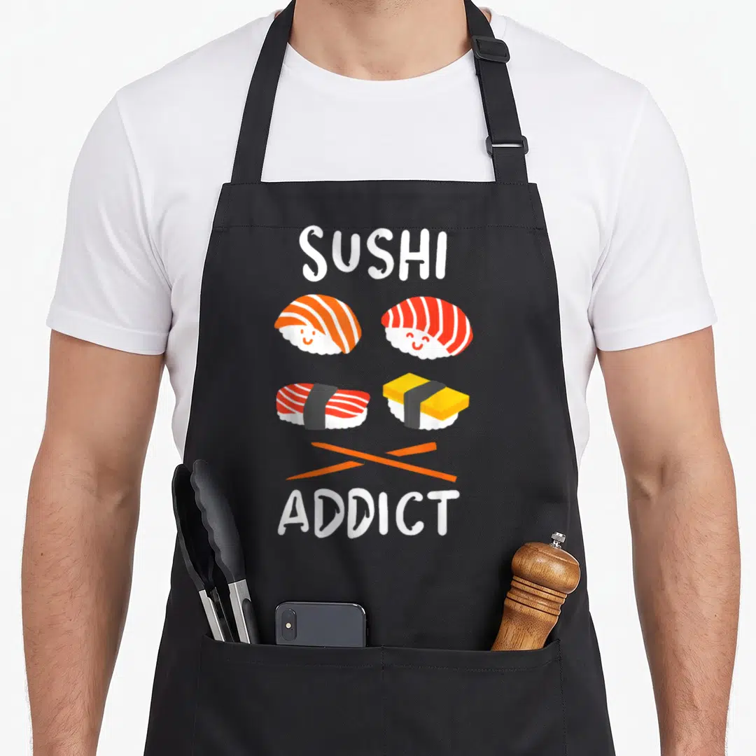 Restaurant sushi apron - SUSHI ADDICT text above four smiling cartoon sushi pieces and crossed orange chopsticks — shown on a black cotton canvas apron against a white background
