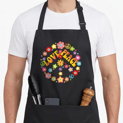 Peace tie apron - Arched LOVE PEACE text with colorful flowers and peace signs in yellow pink blue red green — shown on a black cotton canvas apron against a white background