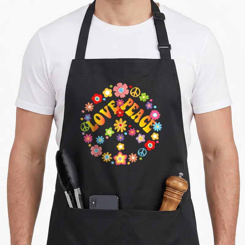 Peace tie apron - Arched LOVE PEACE text with colorful flowers and peace signs in yellow pink blue red green — shown on a black cotton canvas apron against a white background