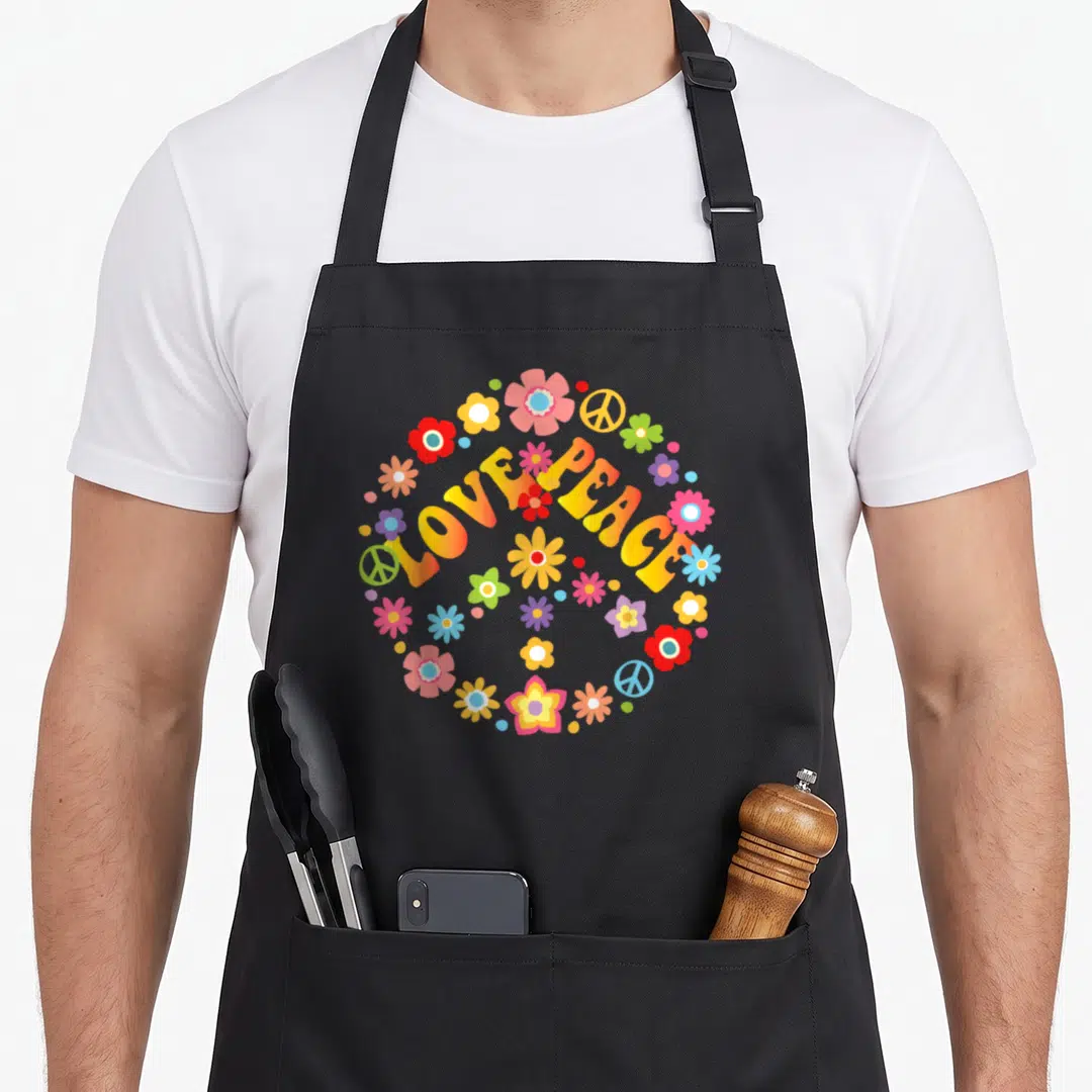 Peace tie apron - Arched LOVE PEACE text with colorful flowers and peace signs in yellow pink blue red green — shown on a black cotton canvas apron against a white background