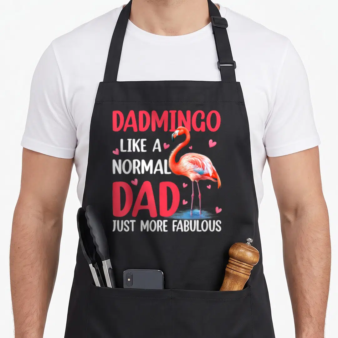 Dadmingo father day apron - Cartoon flamingo in pink and blue overlapping stacked text reading DADMINGO LIKE A NORMAL — shown on a black cotton canvas apron against a white background