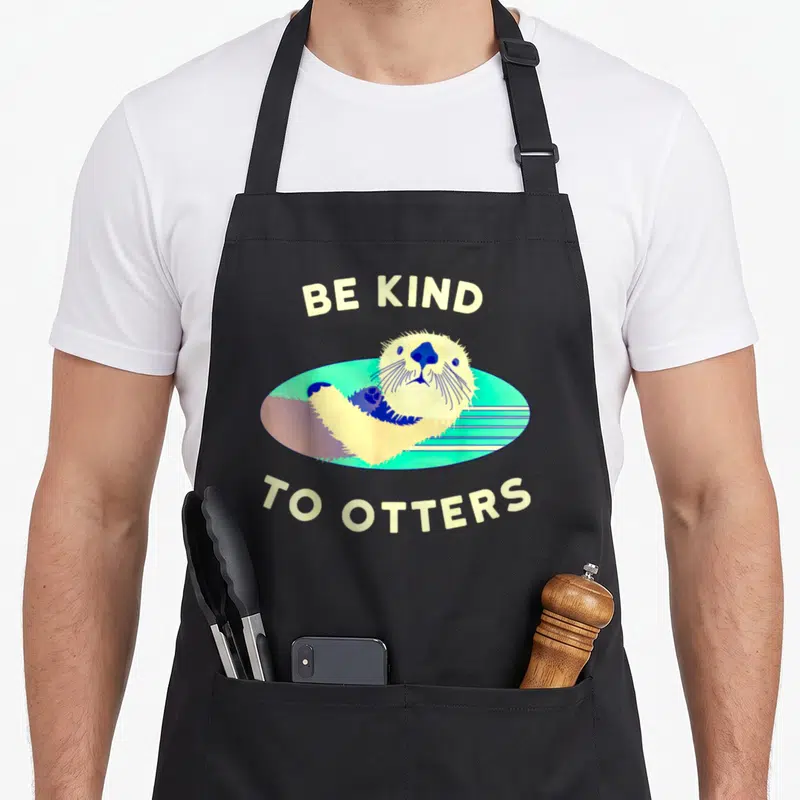 Pun otter lady apron - Cartoon otter floating in cyan oval with arched cream text BE KIND TO OTTERS — shown on a black cotton canvas apron against a white background