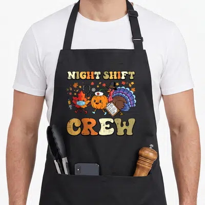 Night thanksgiving apron - NIGHT SHIFT CREW text with cartoon turkey, pumpkin, and leaf in medical gear and pilgrim hat — shown on a black cotton canvas apron against a white background