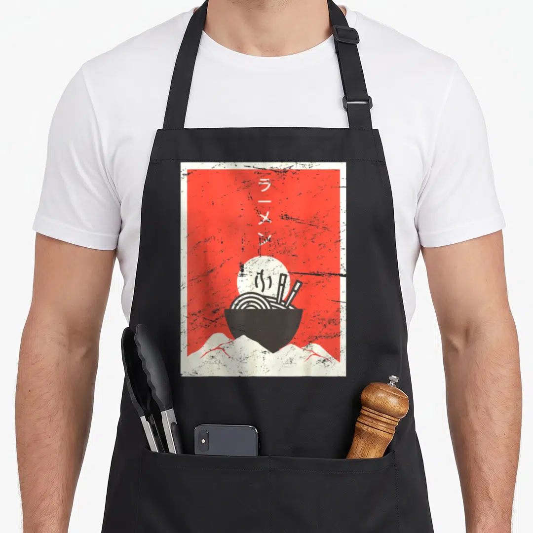 Japanese ramen anime apron - Red poster with black ramen bowl, white sun, mountains, and vertical Japanese text — shown on a black cotton canvas apron against a white background