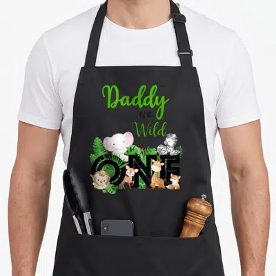 Zoo daddy apron - Cartoon animals and green text reading Daddy Of the Wild ONE clustered around large black letters — shown on a black cotton canvas apron against a white background