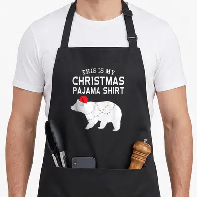 Bear family christmas aprons - Polar bear in red hat wrapped in colored lights under stacked white text — shown on a black cotton canvas apron against a white background