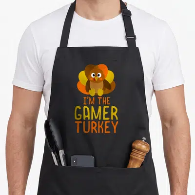 Gamer turkey thanksgiving apron - Cartoon brown and orange turkey above three lines of text I'M THE GAMER TURKEY — shown on a black cotton canvas apron against a white background