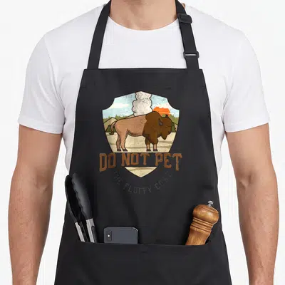 National bison lover apron - Retro shield graphic with brown bison and text Do Not Pet The Fluffy Cows — shown on a black cotton canvas apron against a white background