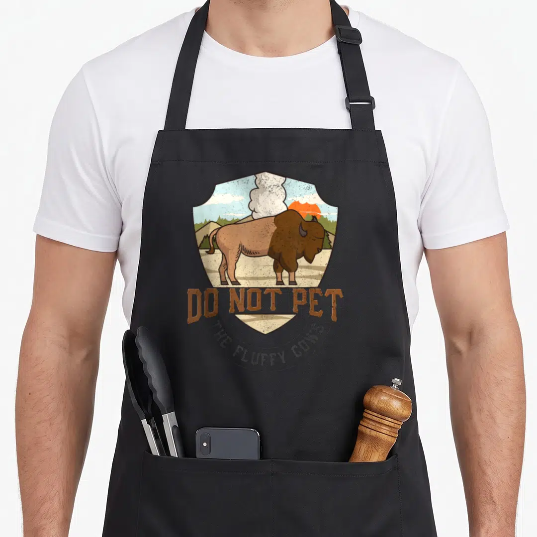 National bison lover apron - Retro shield graphic with brown bison and text Do Not Pet The Fluffy Cows — shown on a black cotton canvas apron against a white background