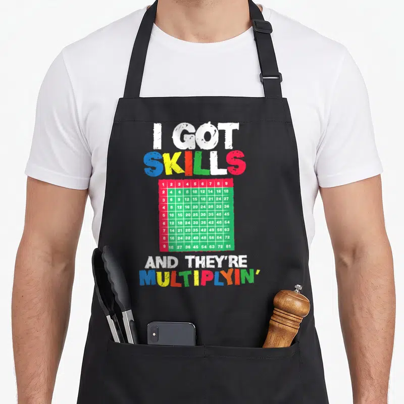 Multiplying teacher apron - I Got Skills And They're Multiplyin' text above a red and green number grid — shown on a black cotton canvas apron against a white background