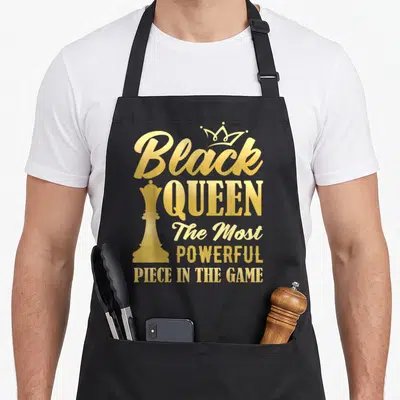 Powerful queen apron - Gold typography reading Black Queen The Most Powerful Piece in The Game with chess piece — shown on a black cotton canvas apron against a white background