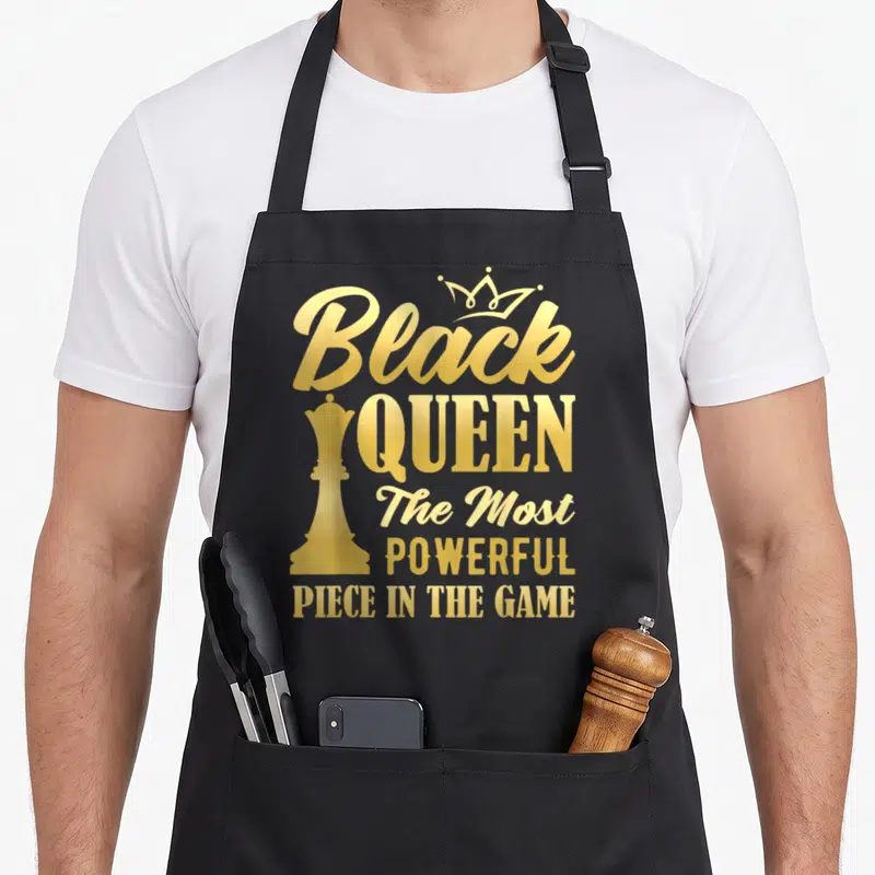 Powerful queen apron - Gold typography reading Black Queen The Most Powerful Piece in The Game with chess piece — shown on a black cotton canvas apron against a white background