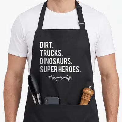 Dirt truck apron - White stacked text reading DIRT TRUCKS DINOSAURS SUPER HEROES #boymomlife on black background — shown on a black cotton canvas apron against a white background