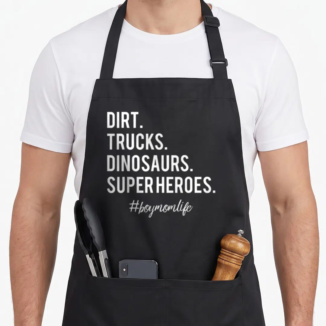 Dirt truck apron - White stacked text reading DIRT TRUCKS DINOSAURS SUPER HEROES #boymomlife on black background — shown on a black cotton canvas apron against a white background