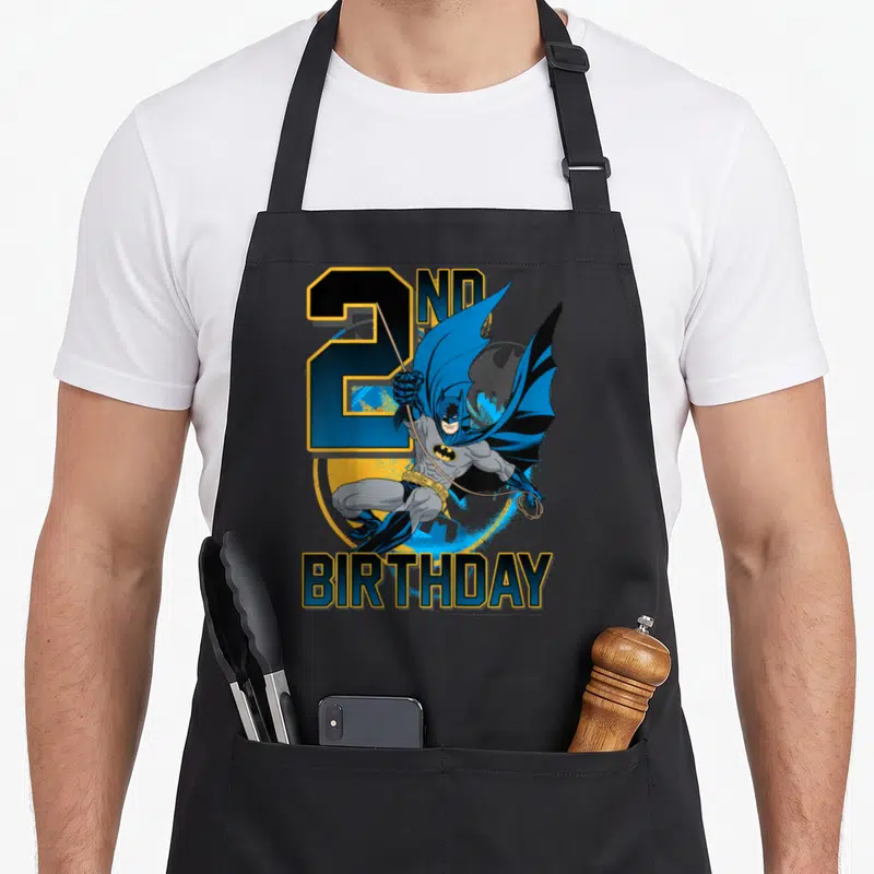 Action 2nd apron - Leaping Batman over large number 2 and yellow bat signal with 2nd Birthday text in blue and grey — shown on a black cotton canvas apron against a white background