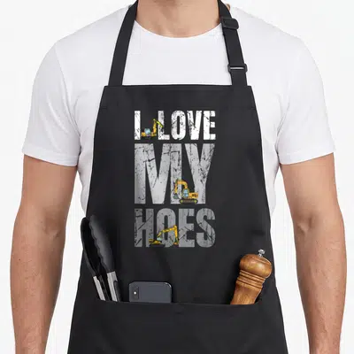 Pun construction apron - Three yellow excavators overlapped by white text reading I LOVE MY HOES on black background — shown on a black cotton canvas apron against a white background