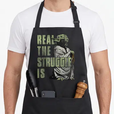 Struggle yoda apron - Yoda holding cane next to stacked green text Real The Struggle Is distressed print — shown on a black cotton canvas apron against a white background