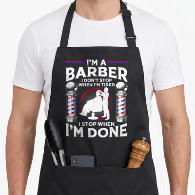 Funny barber hairstyling apron - I'M A BARBER typography with vintage pole graphics and stylist silhouette — shown on a black cotton canvas apron against a white background
