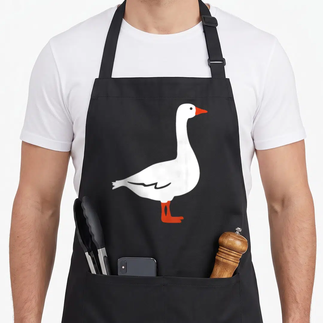 Funny goose apron - Minimalist white goose with orange beak and legs facing right — shown on a black cotton canvas apron against a white background