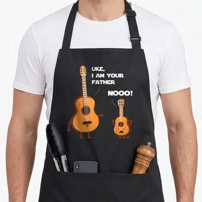 Uke ukulele apron - Orange cartoon guitar and ukulele with white text UKE I AM YOUR FATHER NOOO — shown on a black cotton canvas apron against a white background