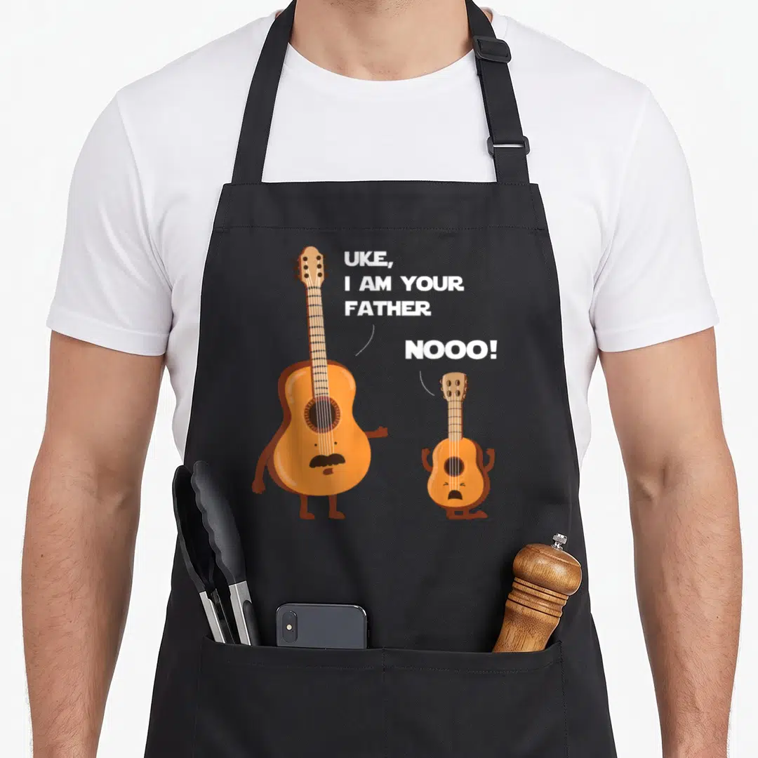 Uke ukulele apron - Orange cartoon guitar and ukulele with white text UKE I AM YOUR FATHER NOOO — shown on a black cotton canvas apron against a white background