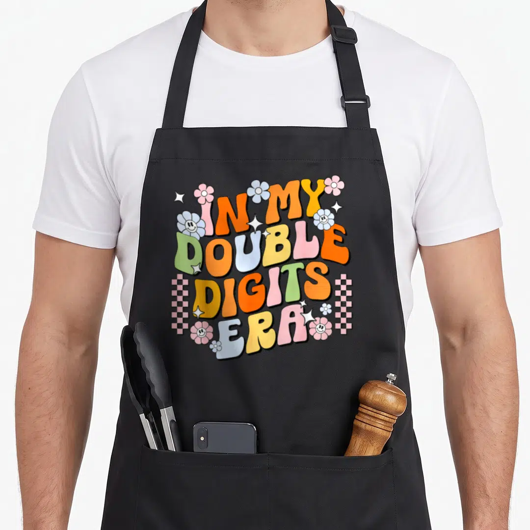 Digit era birthday apron - IN MY DOUBLE DIGITS ERA text in orange yellow blue green pink bubbly font with flowers — shown on a black cotton canvas apron against a white background