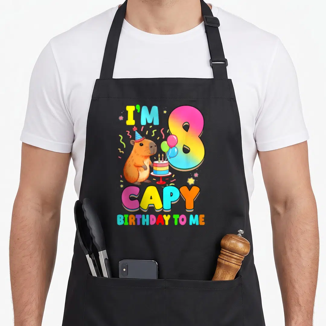 8th capybara birthday apron - Cartoon capybara with party hat, rainbow number 8, and cake surrounded by confetti — shown on a black cotton canvas apron against a white background
