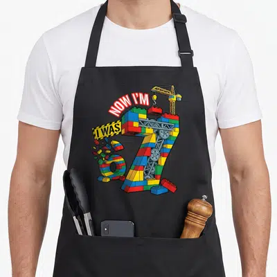 Funny brick building aprons - Number 7 made of toy bricks with crane and caution strip text — shown on a black cotton canvas apron against a white background