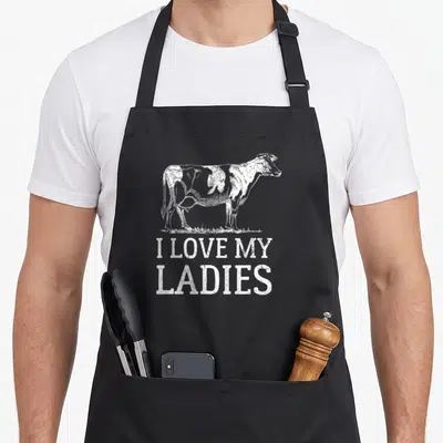90s cow print aprons - Distressed white cow standing on grass above I Love My Ladies text — shown on a black cotton canvas apron against a white background