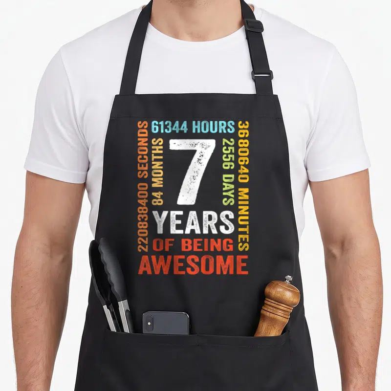 7th month birthday apron - White number 7 surrounded by orange blue and green time stats and years of being awesome text — shown on a black cotton canvas apron against a white background