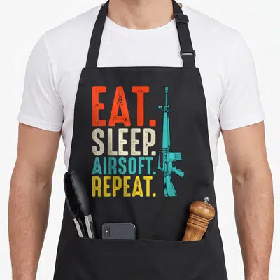 Eat sports team aprons - Stacked EAT SLEEP AIRSOFT REPEAT text in red beige blue yellow next to a light blue airsoft — shown on a black cotton canvas apron against a white background