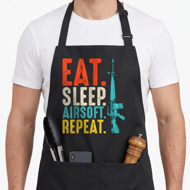 Eat sports team aprons - Stacked EAT SLEEP AIRSOFT REPEAT text in red beige blue yellow next to a light blue airsoft — shown on a black cotton canvas apron against a white background