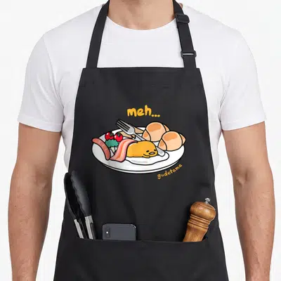 Meh gudetama apron - Yellow egg character on white plate with bacon, sausage, tomatoes, and meh... text in orange — shown on a black cotton canvas apron against a white background