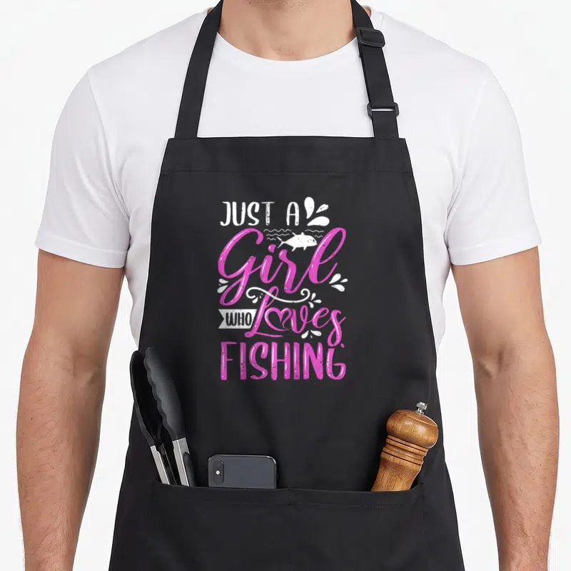 Cute fisher fishing aprons - Pink and white stacked text reading Just A Girl Who Loves Fishing with fish and swirl — shown on a black cotton canvas apron against a white background