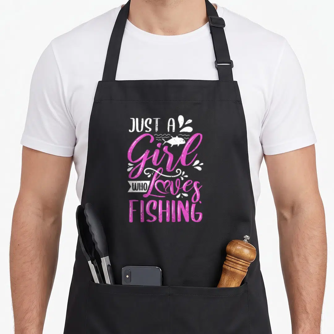 Cute fisher fishing aprons - Pink and white stacked text reading Just A Girl Who Loves Fishing with fish and swirl — shown on a black cotton canvas apron against a white background