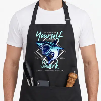 Unless ecologist apron - Blue cartoon shark inside white diamond frame with humorous text about being a shark — shown on a black cotton canvas apron against a white background