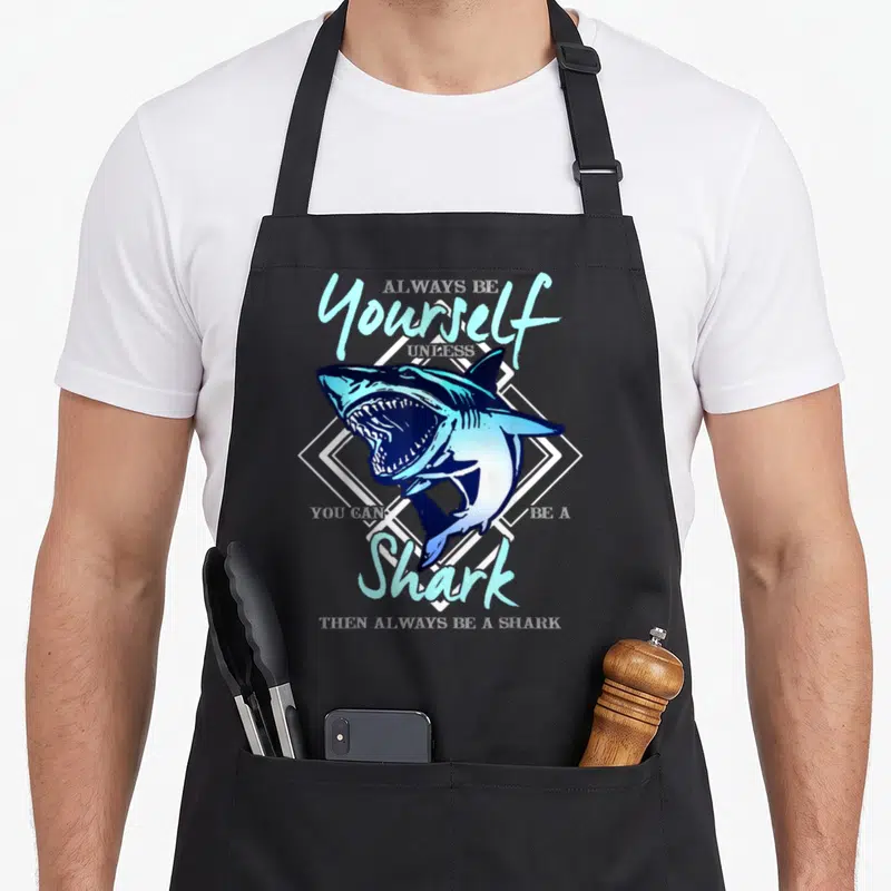 Unless ecologist apron - Blue cartoon shark inside white diamond frame with humorous text about being a shark — shown on a black cotton canvas apron against a white background