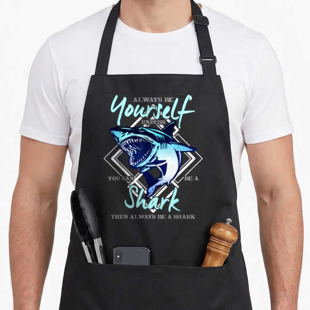 Unless ecologist apron - Blue cartoon shark inside white diamond frame with humorous text about being a shark — shown on a black cotton canvas apron against a white background
