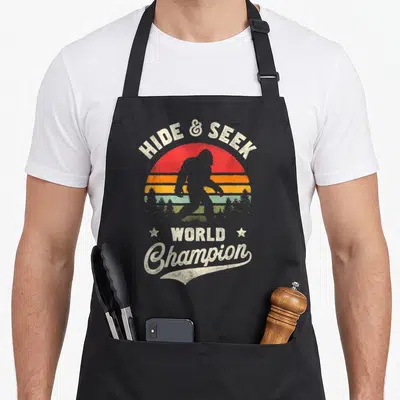 Seek bigfoot apron - Retro sun graphic with bigfoot silhouette and text hide and seek world champion — shown on a black cotton canvas apron against a white background