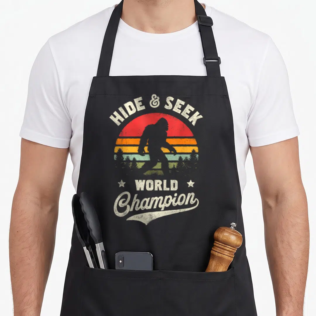 Seek bigfoot apron - Retro sun graphic with bigfoot silhouette and text hide and seek world champion — shown on a black cotton canvas apron against a white background