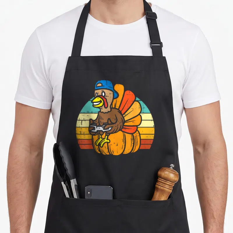 Gamer pumpkin apron - Cartoon turkey in blue cap holding controller on pumpkin with striped sun — shown on a black cotton canvas apron against a white background