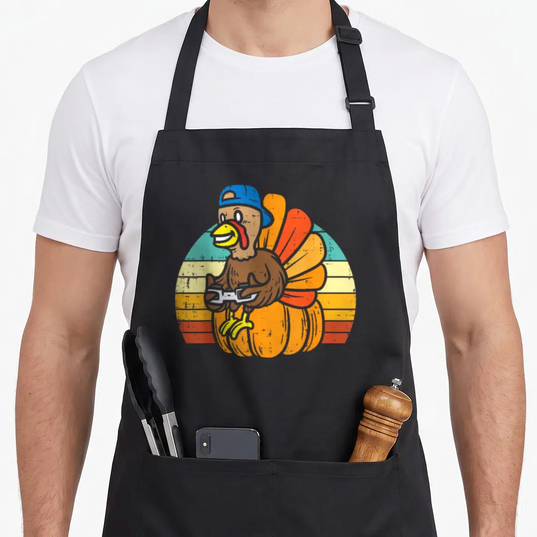 Gamer pumpkin apron - Cartoon turkey in blue cap holding controller on pumpkin with striped sun — shown on a black cotton canvas apron against a white background