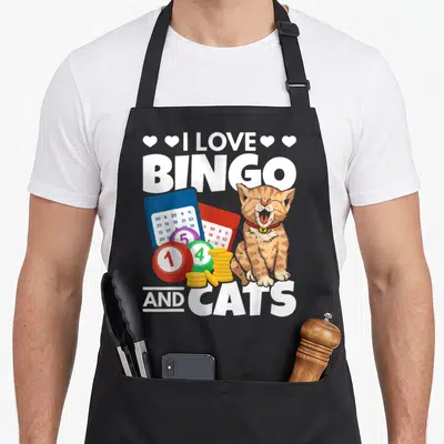 Cat bingo apron - Orange cat sitting next to blue and red bingo cards, colored balls, and gold coin stacks under white — shown on a black cotton canvas apron against a white background
